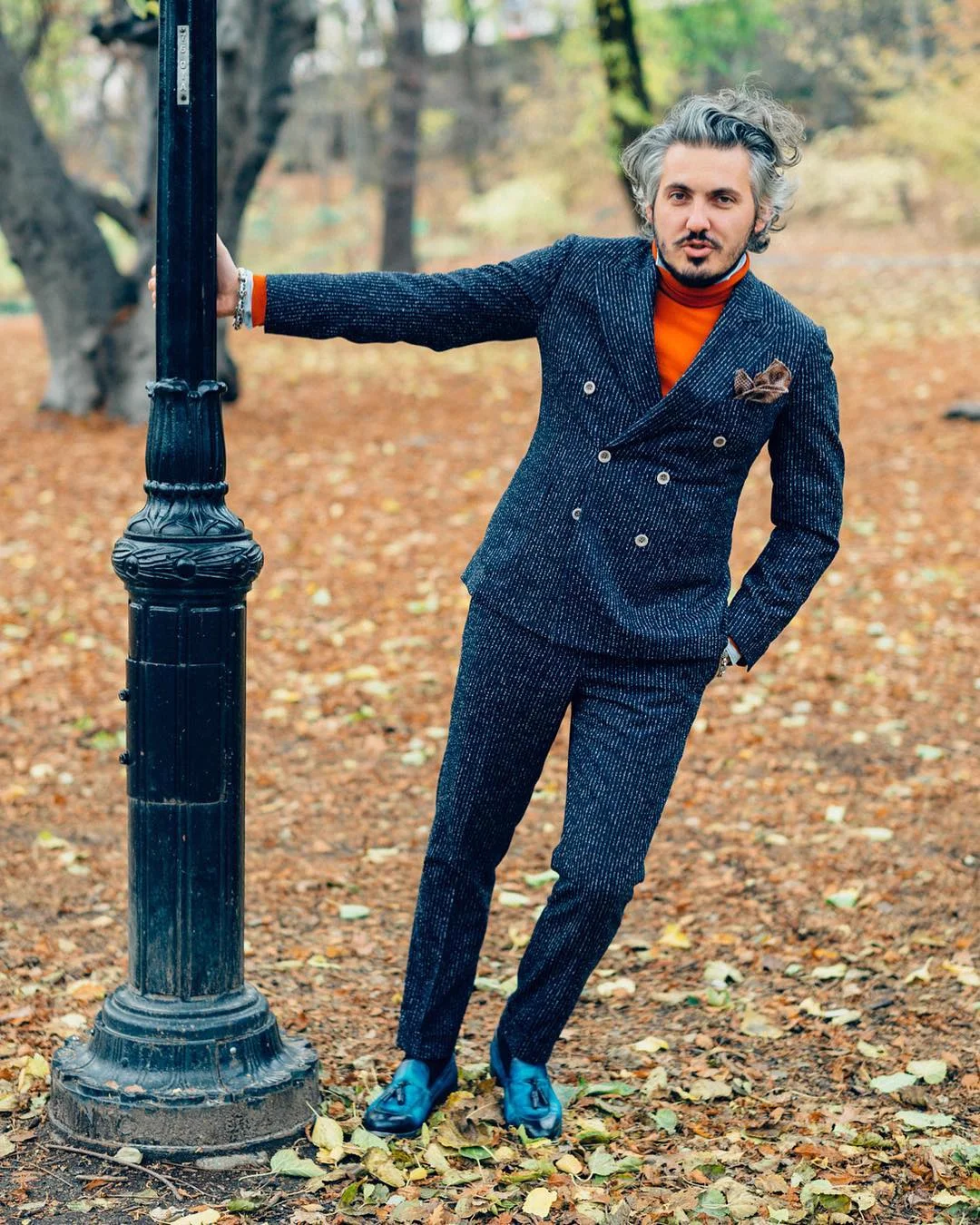 Matteo Perin leaning on a lamppost in an autumn park, in a navy chalk-stripe double-breasted suit over an orange roll-neck