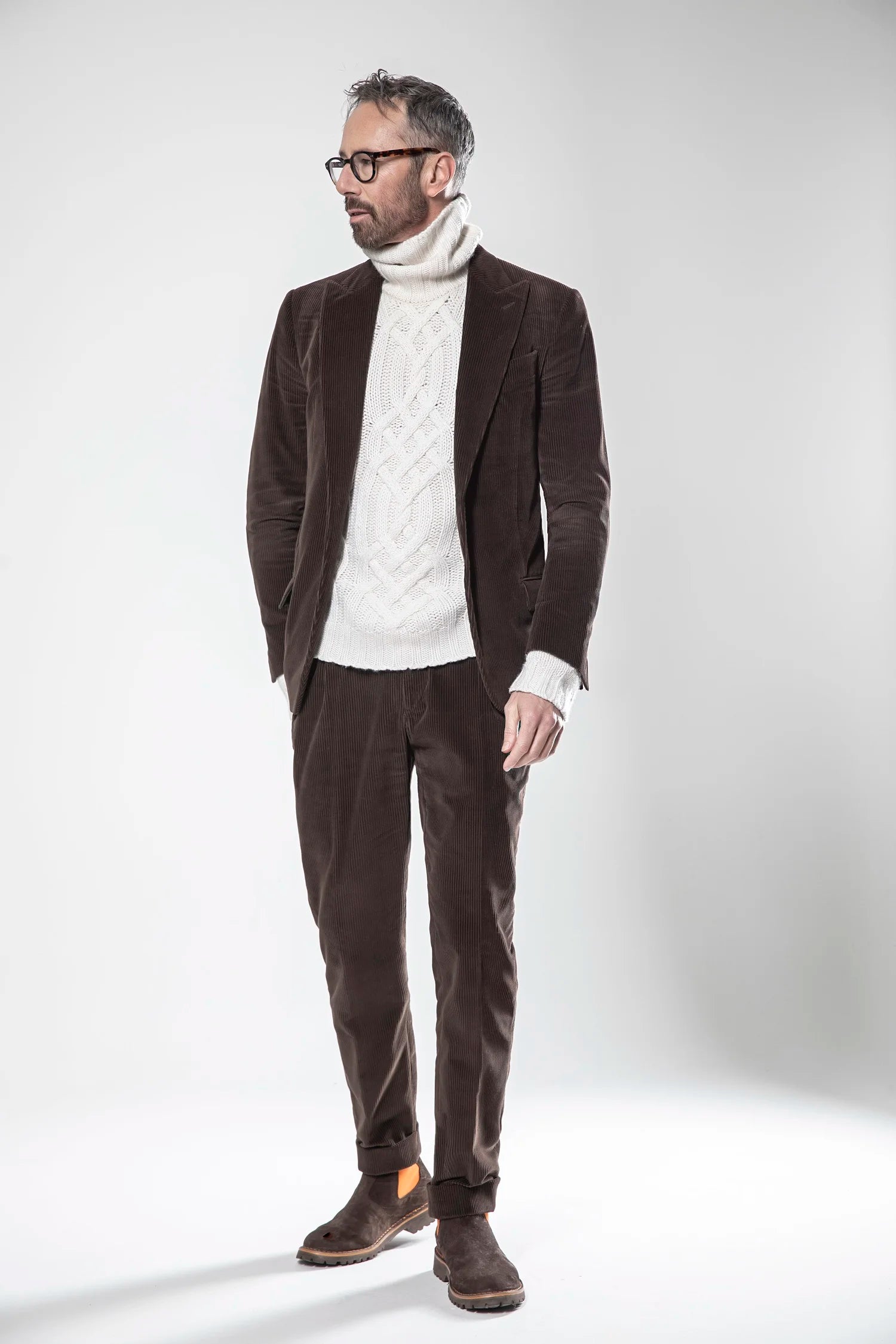 Man wearing a brown blazer, white turtleneck sweater, brown pants, and brown shoes on a white background