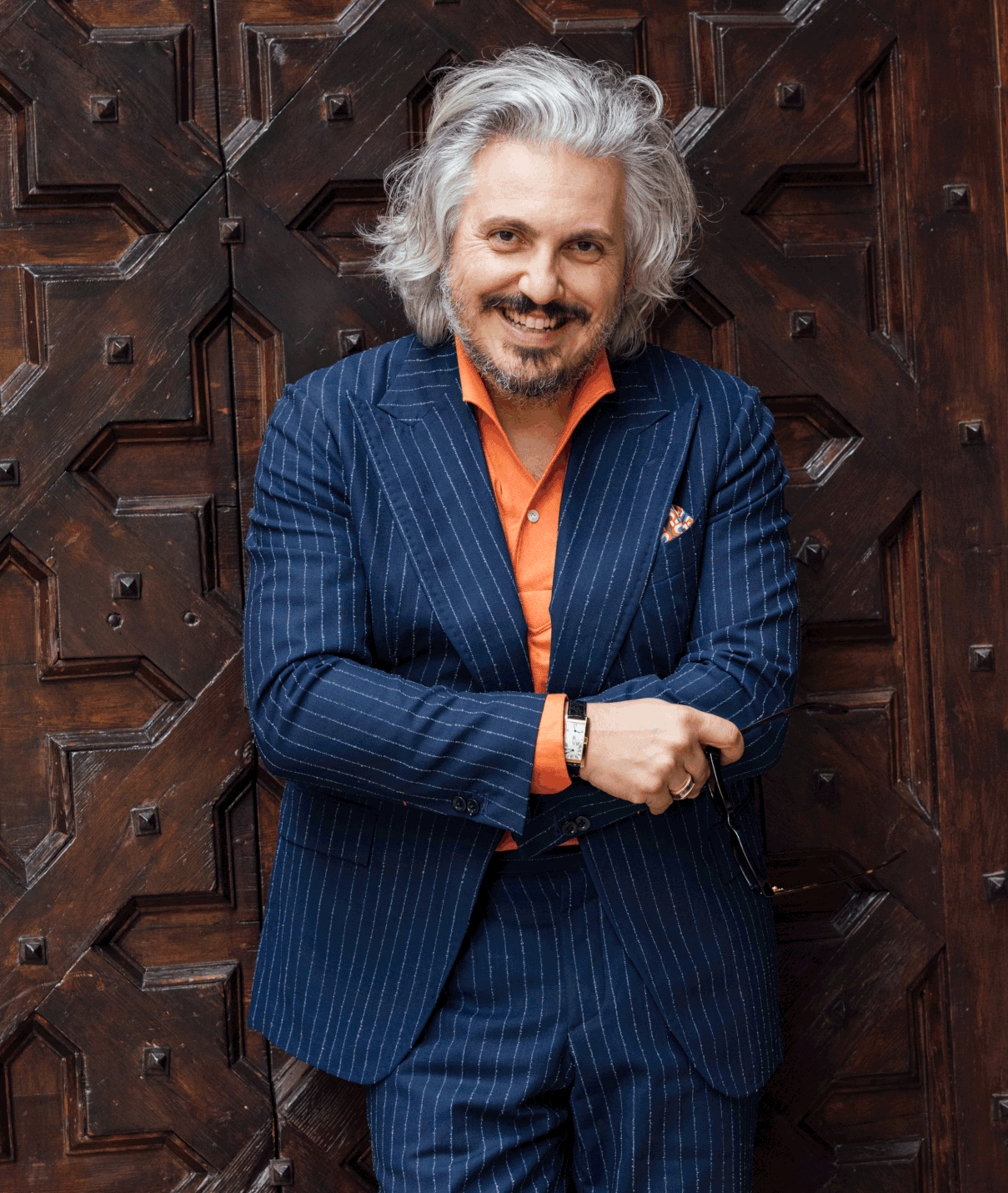 Man with gray hair and beard wearing a blue pinstripe suit and orange shirt stands smiling in front of an ornate wooden door.