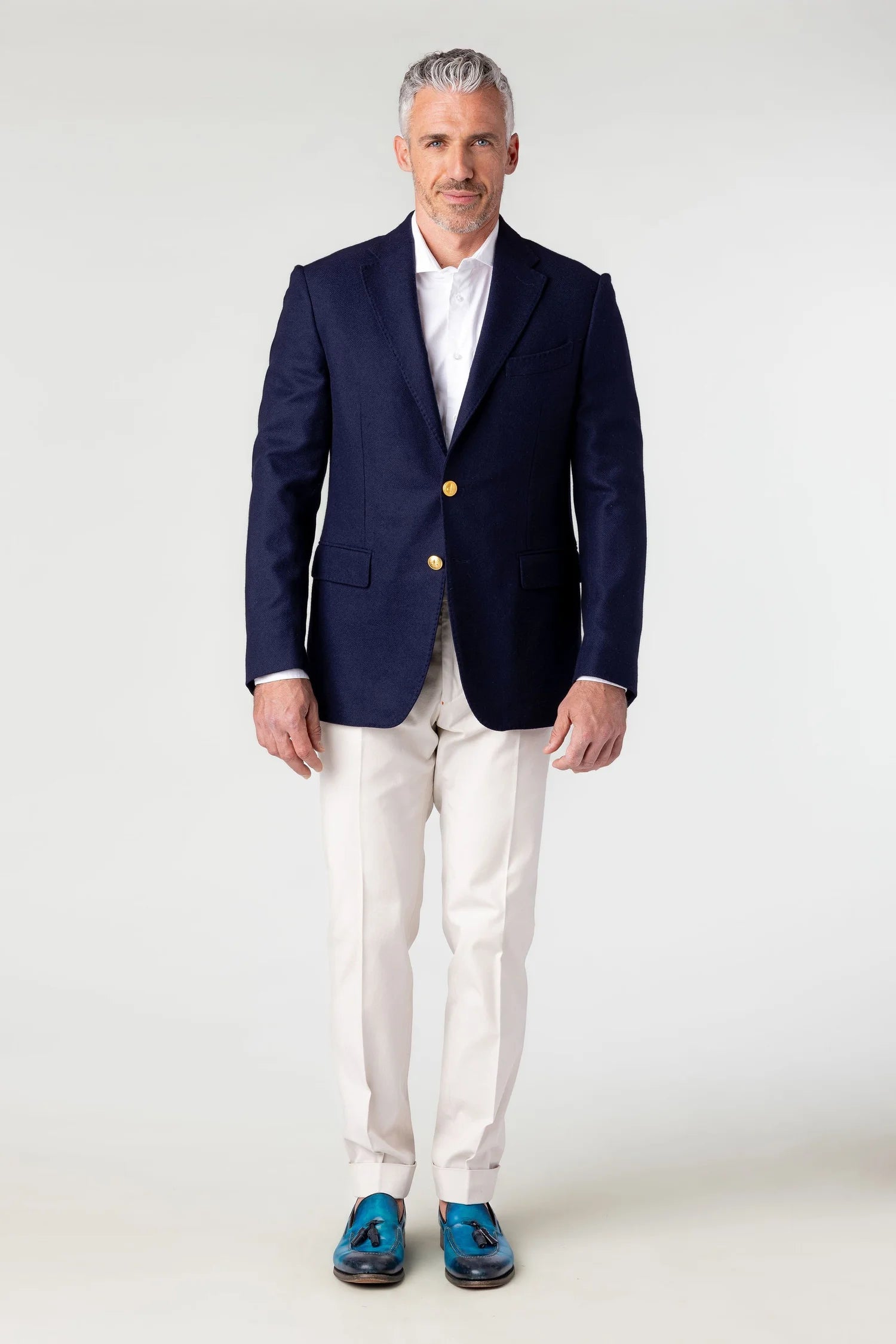 Man wearing a navy blazer with gold buttons, white shirt, and white pants on a white background
