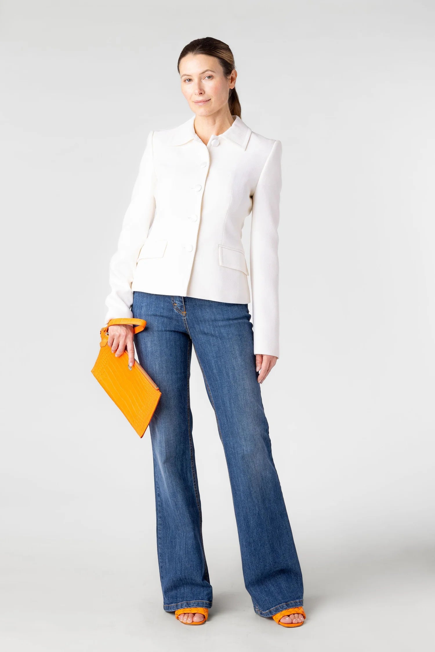 Woman wearing a white jacket and blue jeans holding an orange clutch against a white background