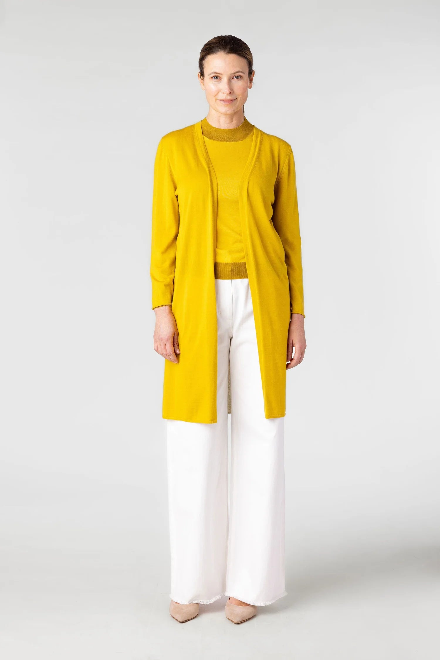 Woman wearing a yellow cardigan over a mustard top with white pants on a light gray background
