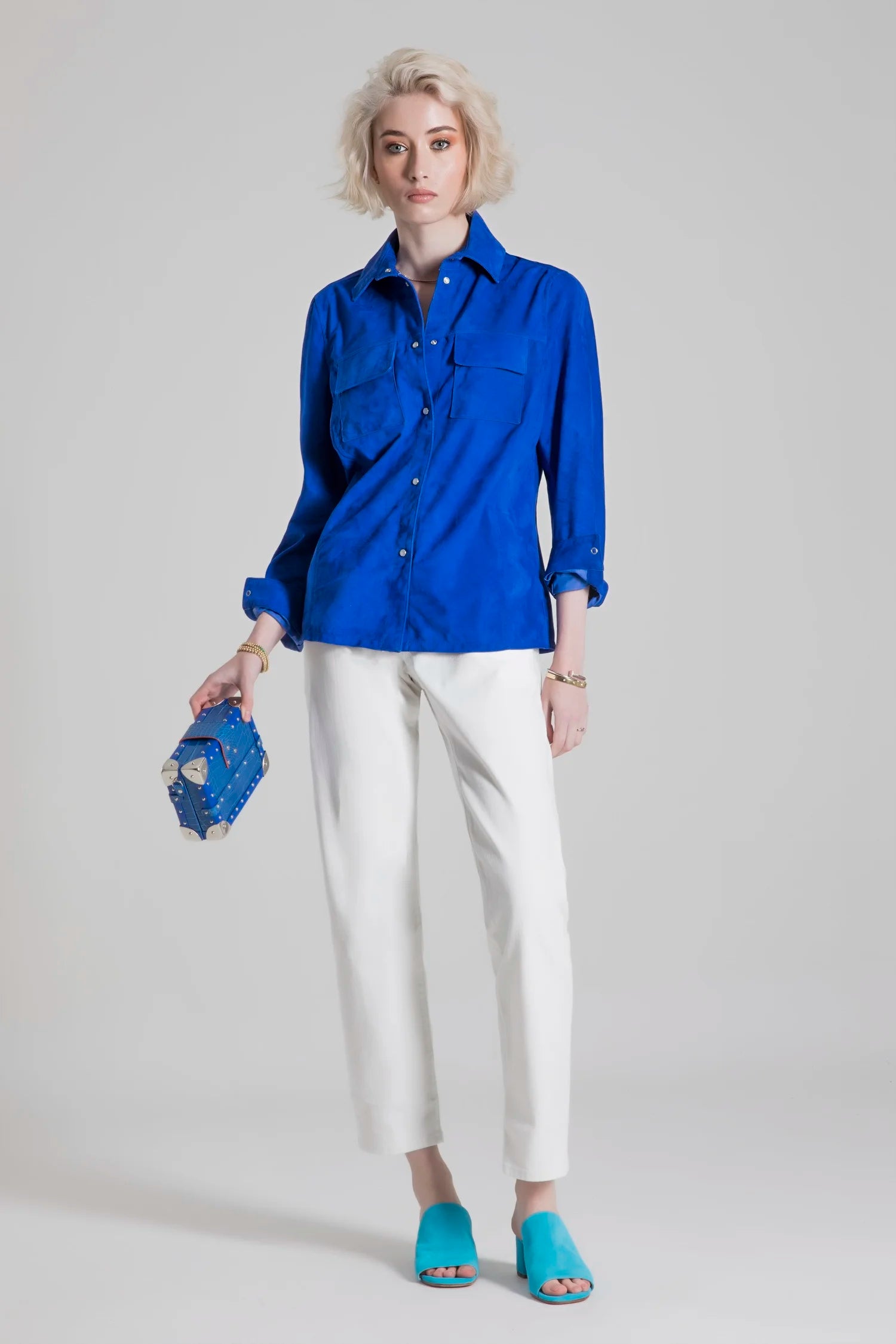 Woman wearing a blue shirt and white pants holding a blue clutch against a gray background