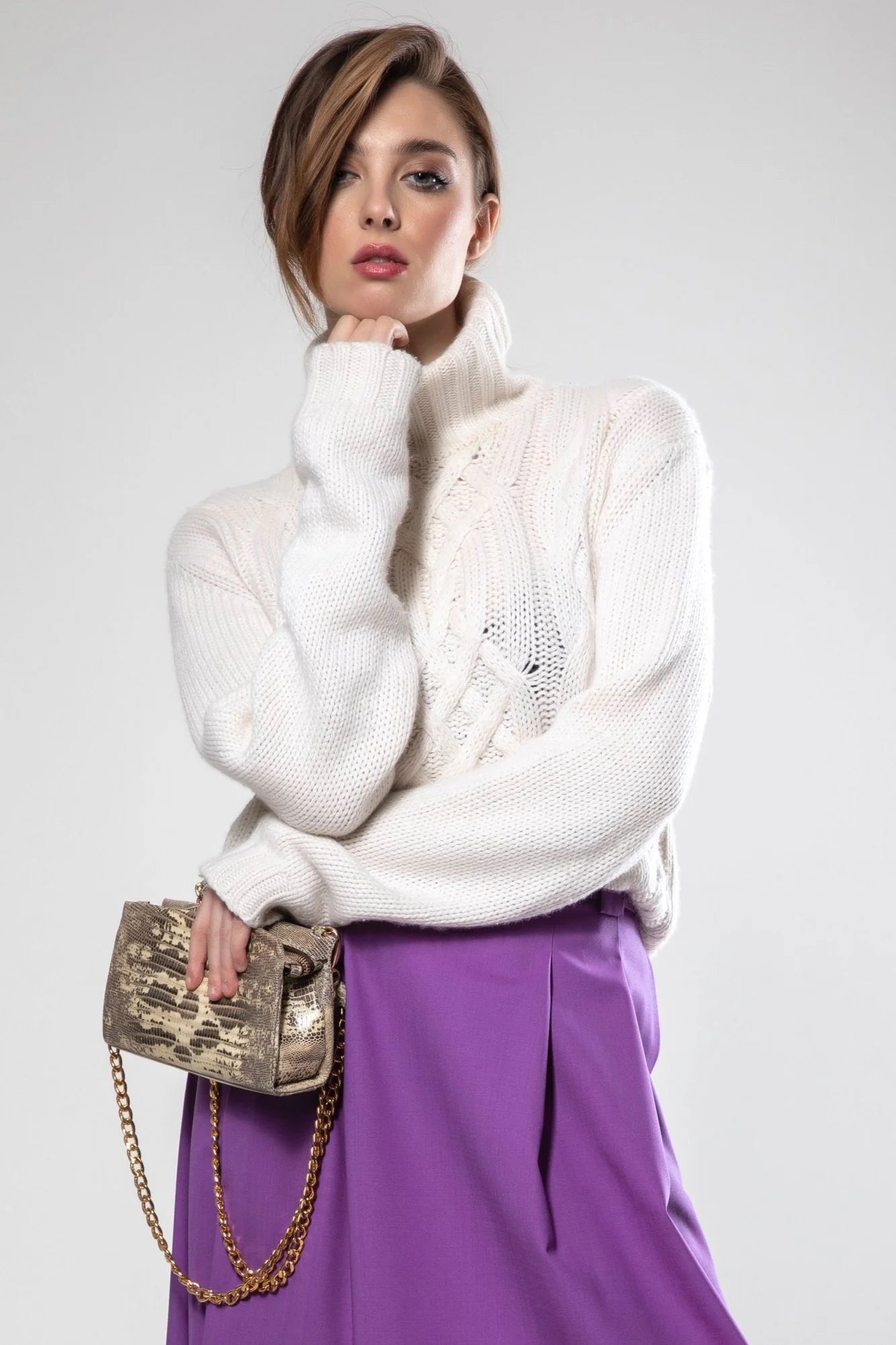 Woman wearing a white sweater and purple skirt with a gold handbag on a light gray background
