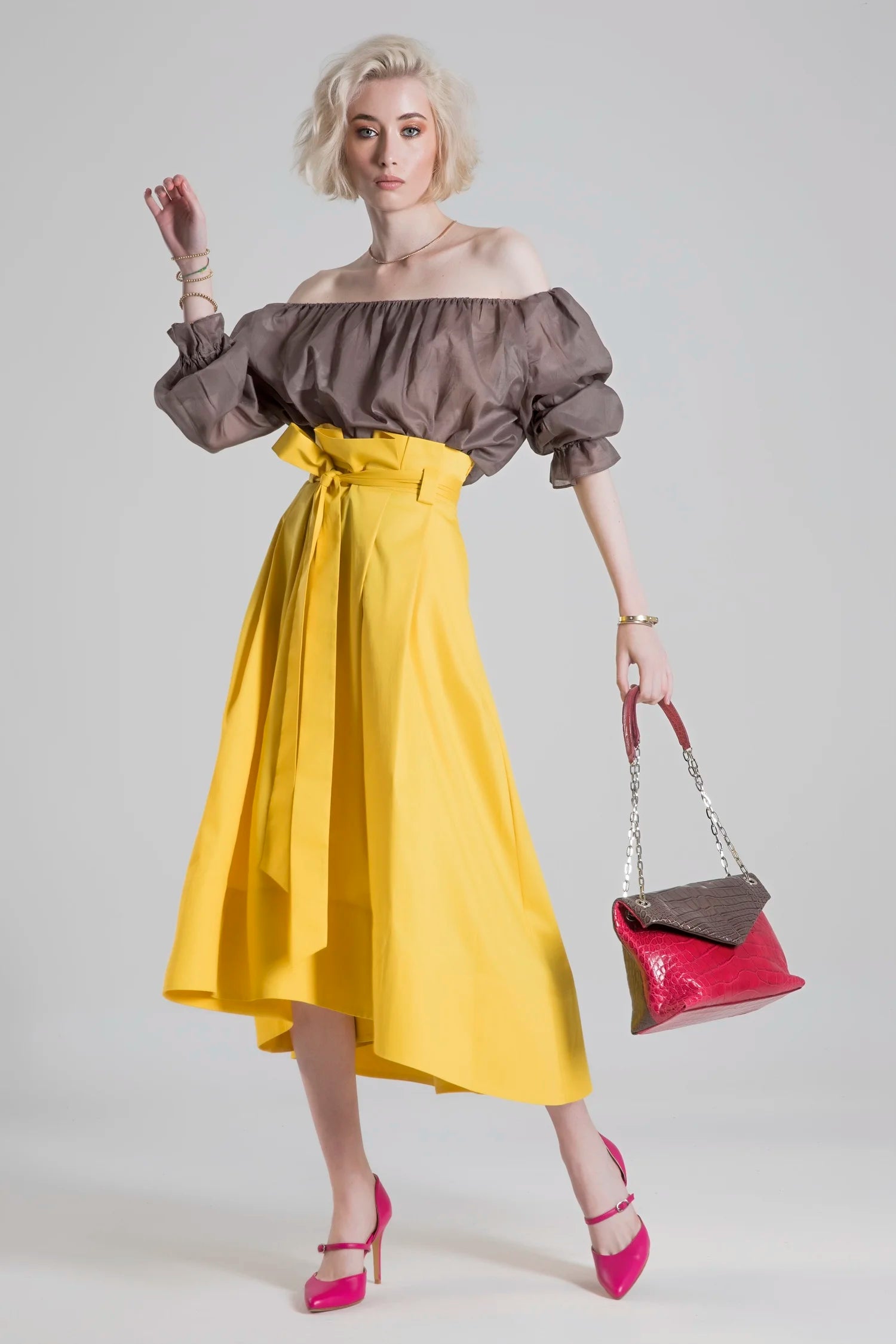 Woman wearing a brown off-shoulder top and yellow skirt, holding a pink handbag on a white background