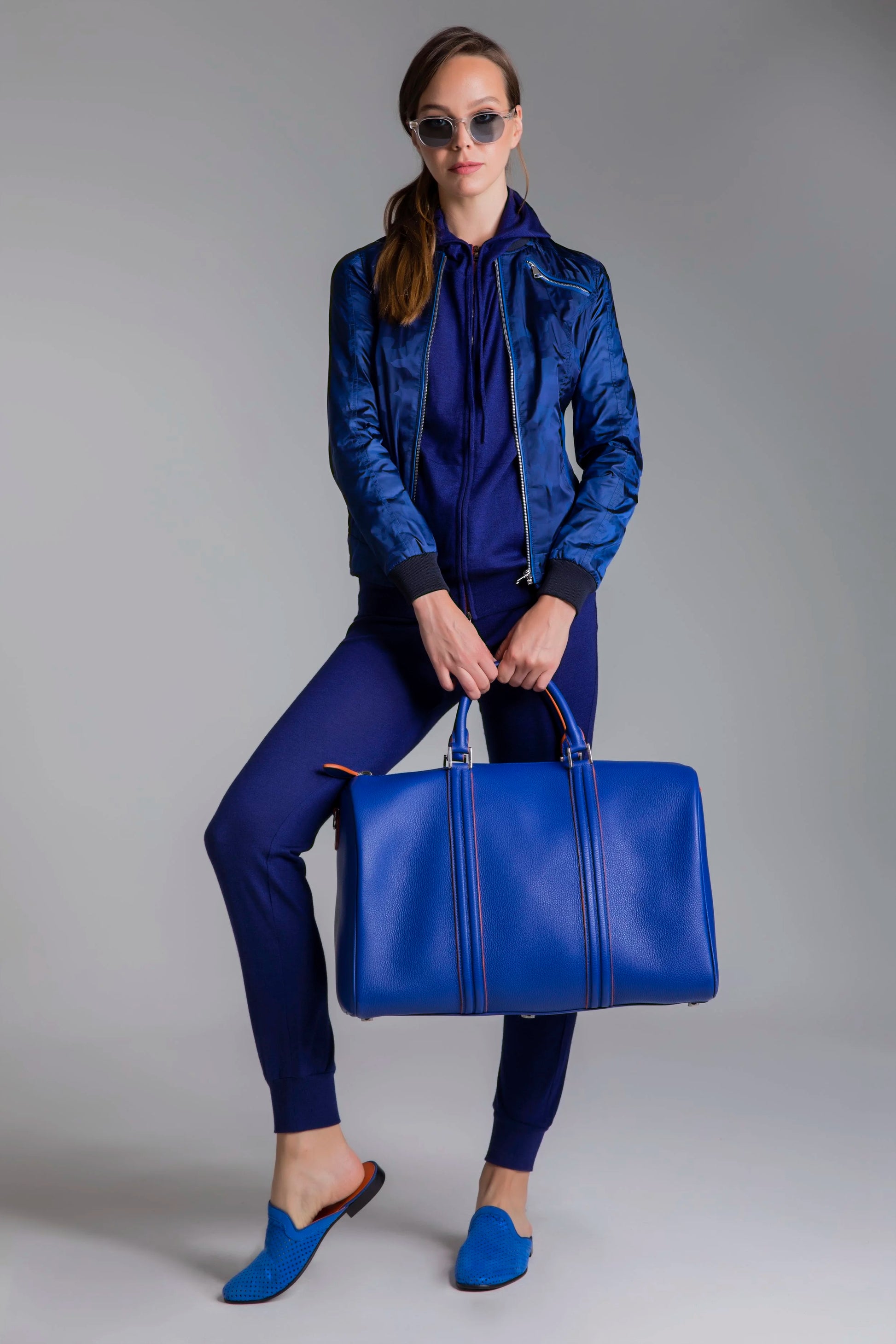 Woman in a blue outfit holding a matching blue bag against a gray background