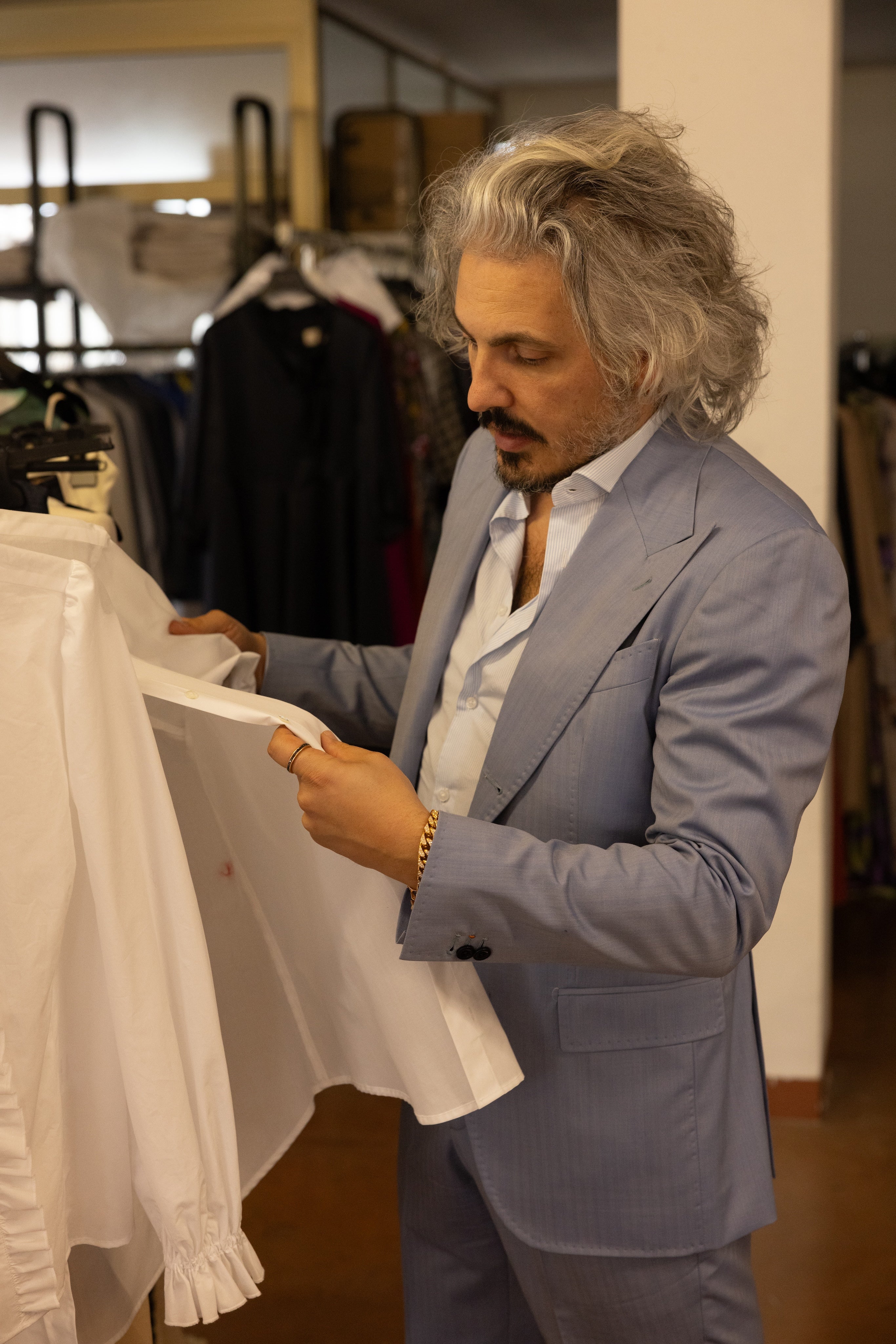 Designer in a light blue suit examining a white dress shirt in a fashion studio, reviewing fabric quality and craftsmanship.
