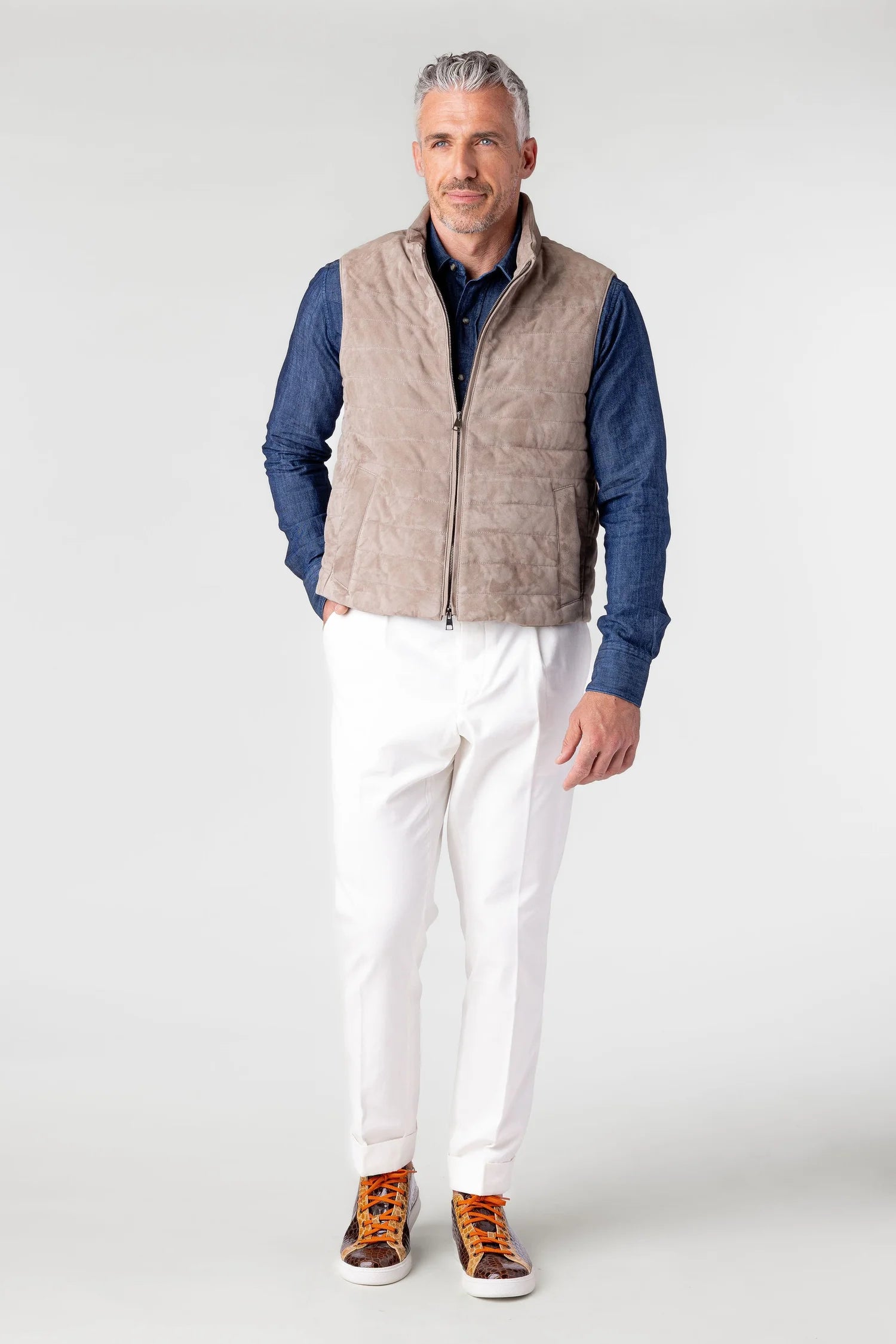 Man wearing a beige quilted vest over a blue shirt with white pants on a white background