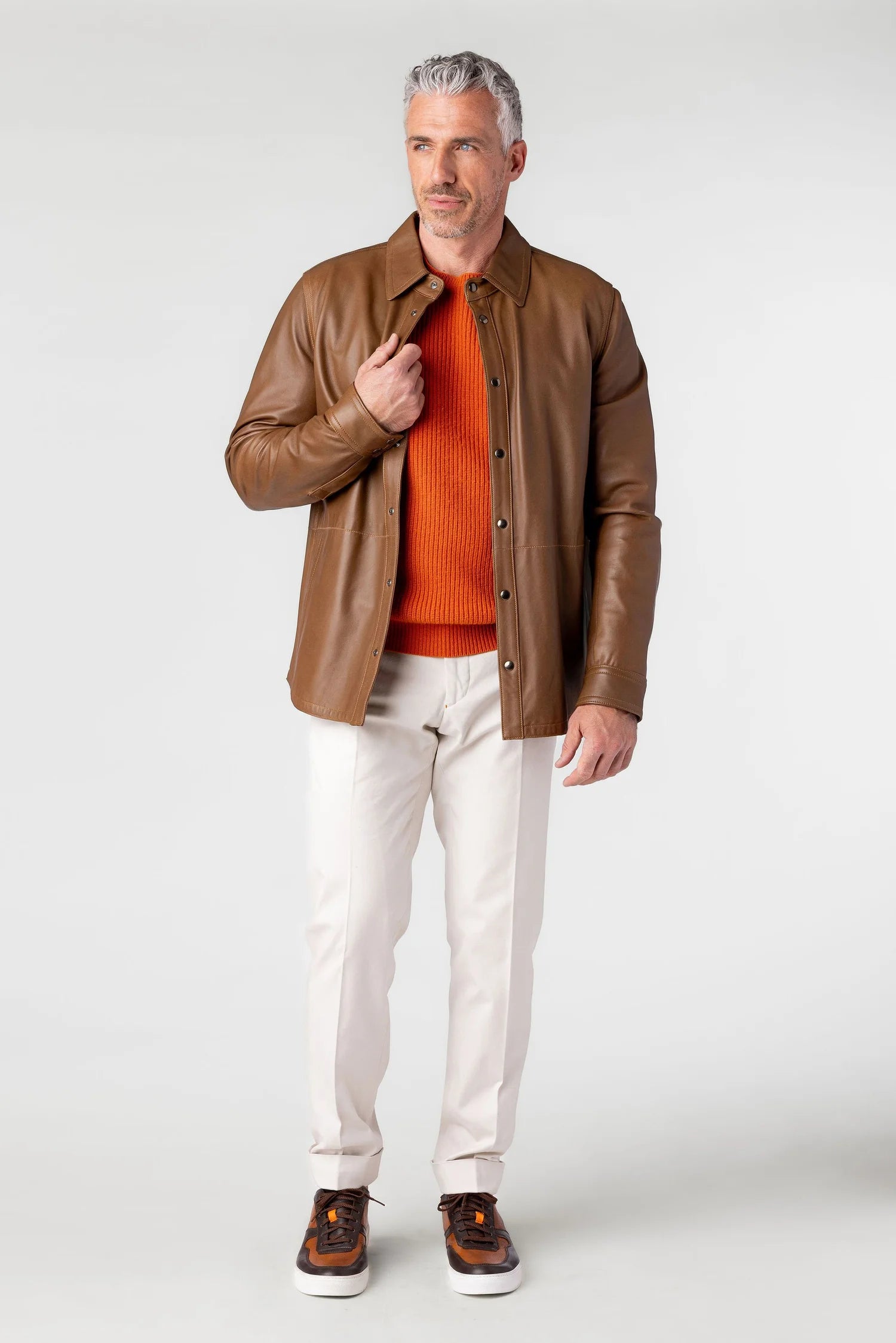 Man wearing a brown leather jacket, orange sweater, and white pants on a white background