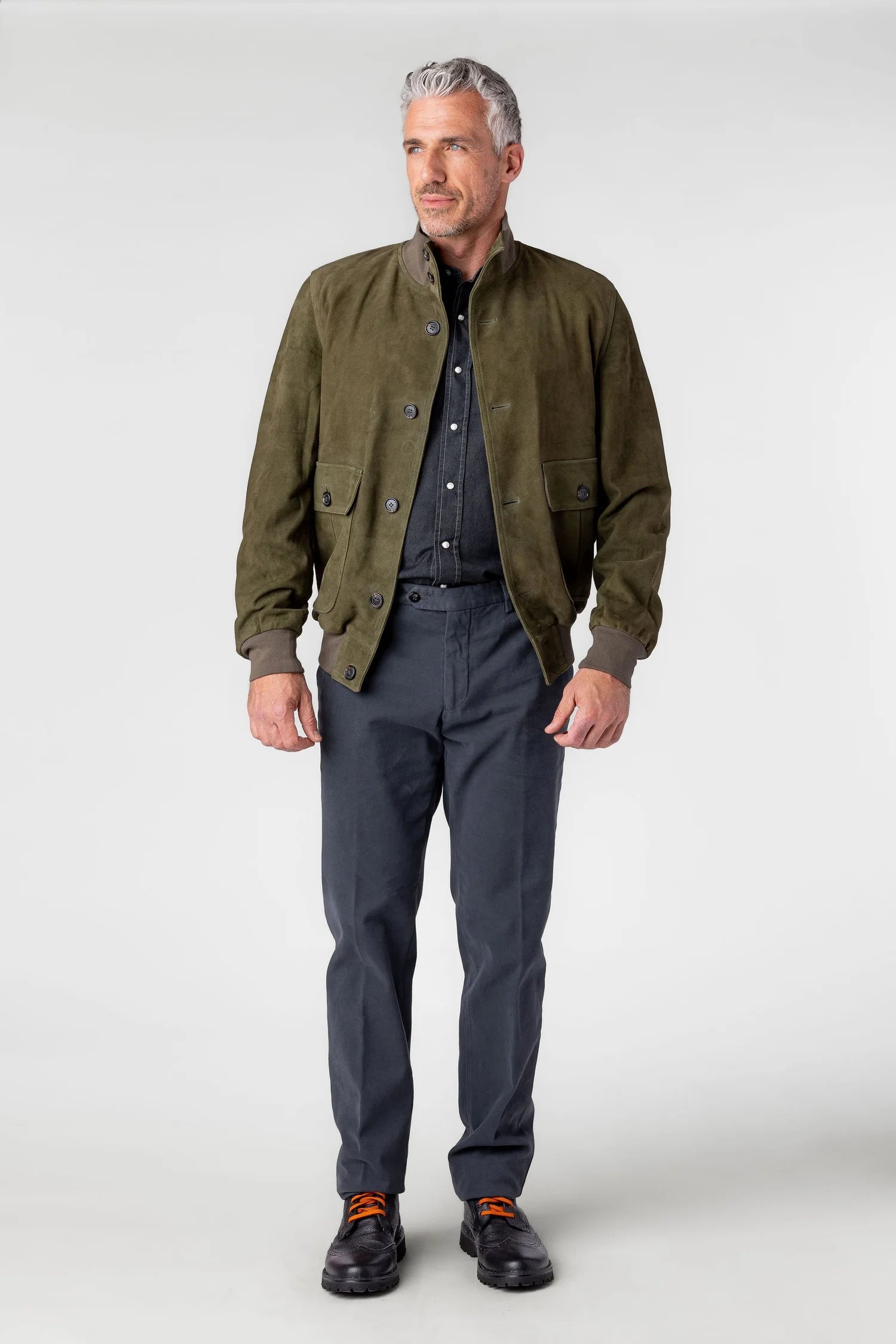 Man wearing an olive green jacket, dark shirt, gray pants, and black shoes on a white background