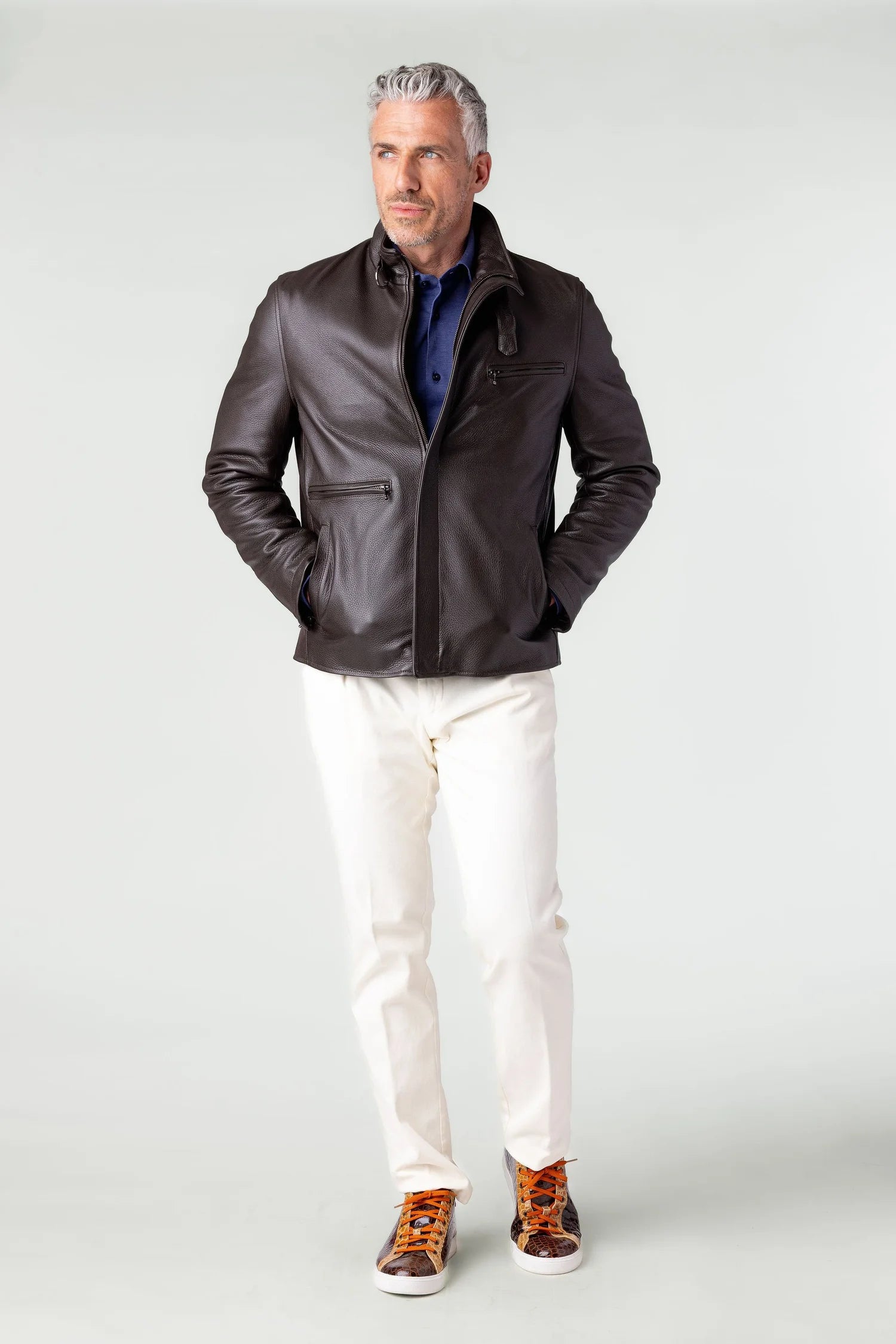 Man wearing a dark leather jacket, white pants, and brown shoes on a light gray background
