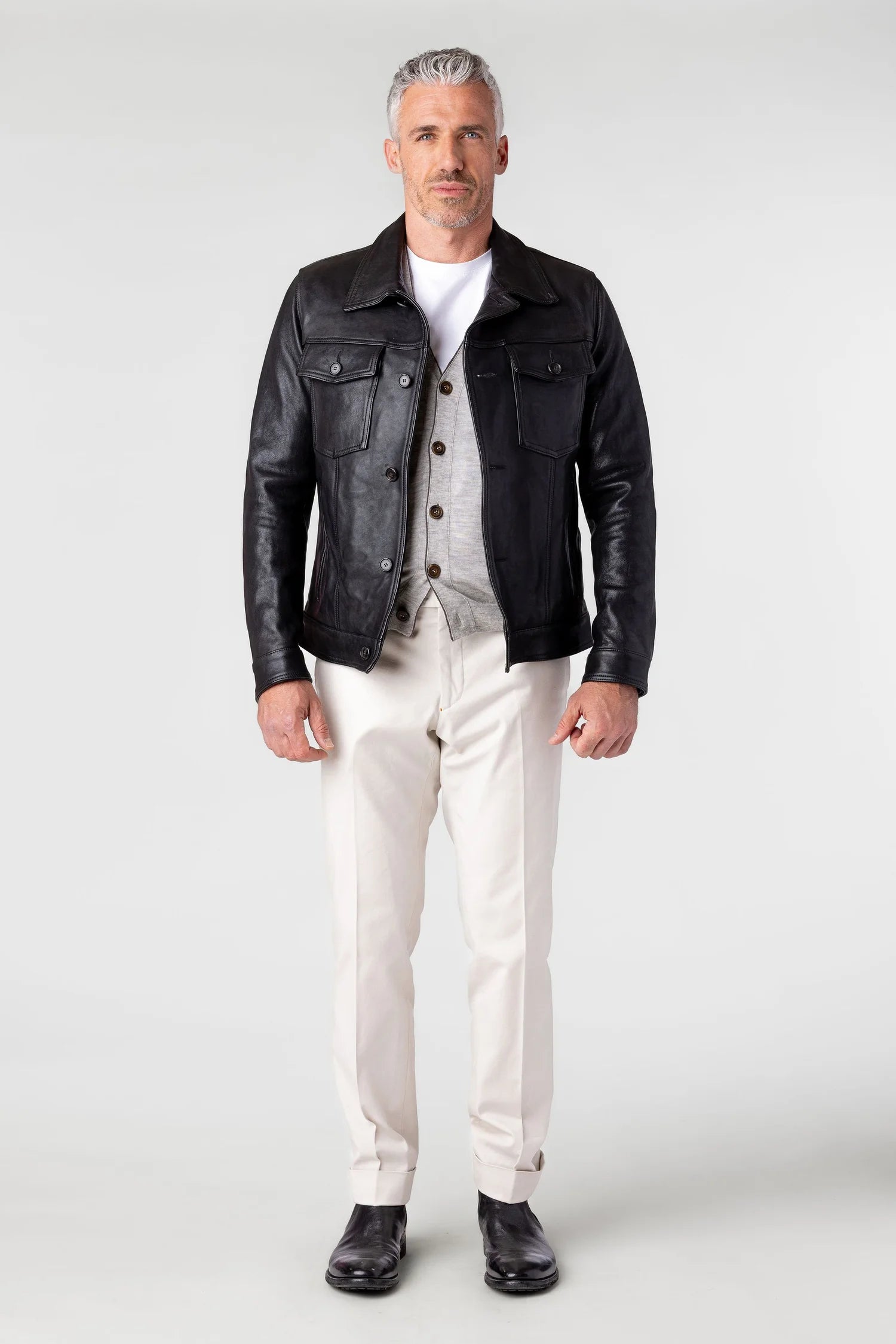 Man wearing a black leather jacket, white shirt, and white pants on a gray background