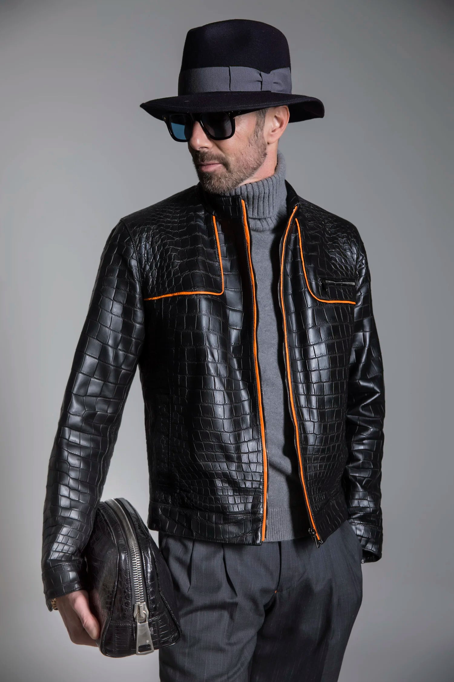 Man wearing a black textured leather jacket with orange accents, holding a black shoe, against a gray background.