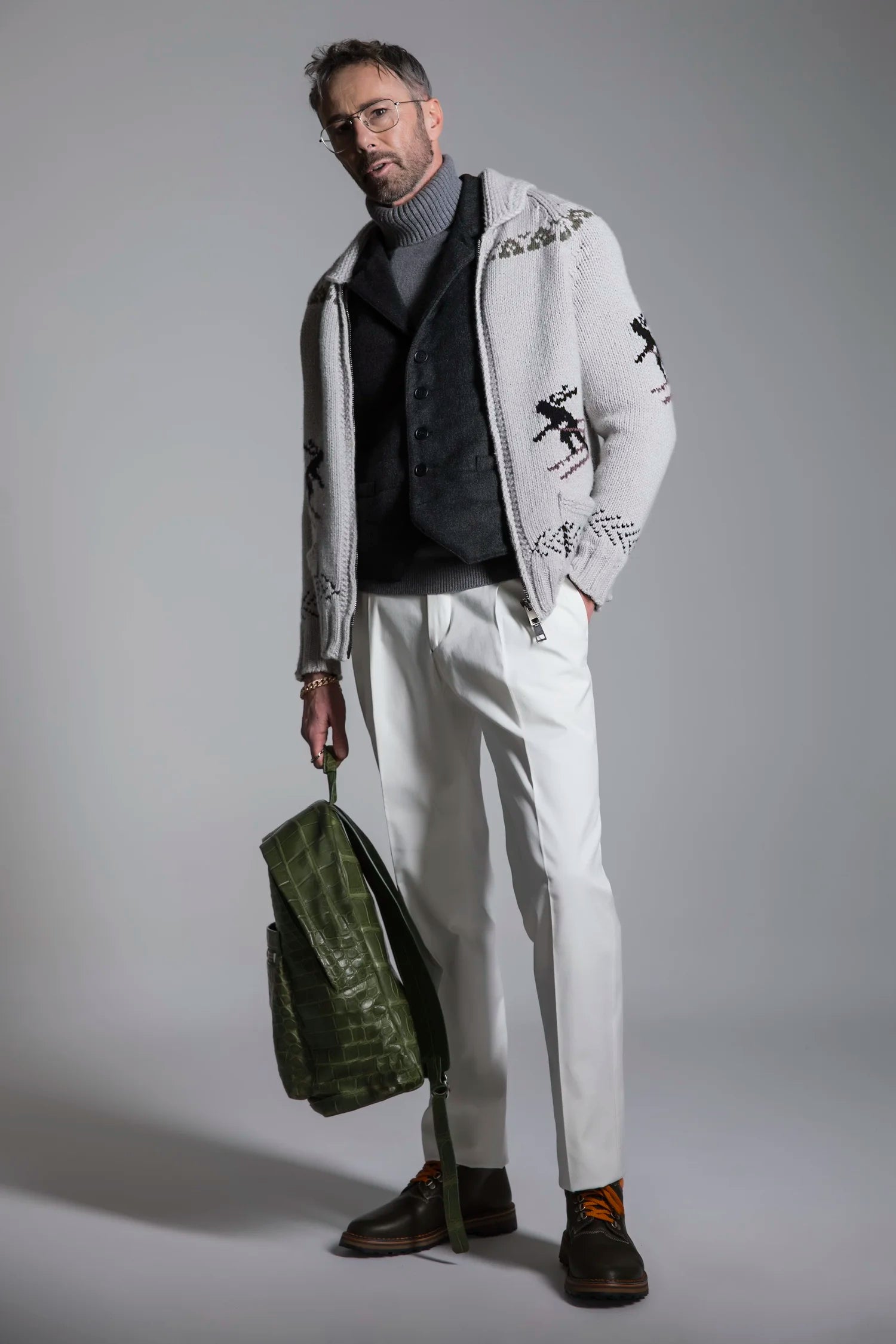 Man wearing a stylish outfit with a white embroidered jacket, holding a green bag against a gray background