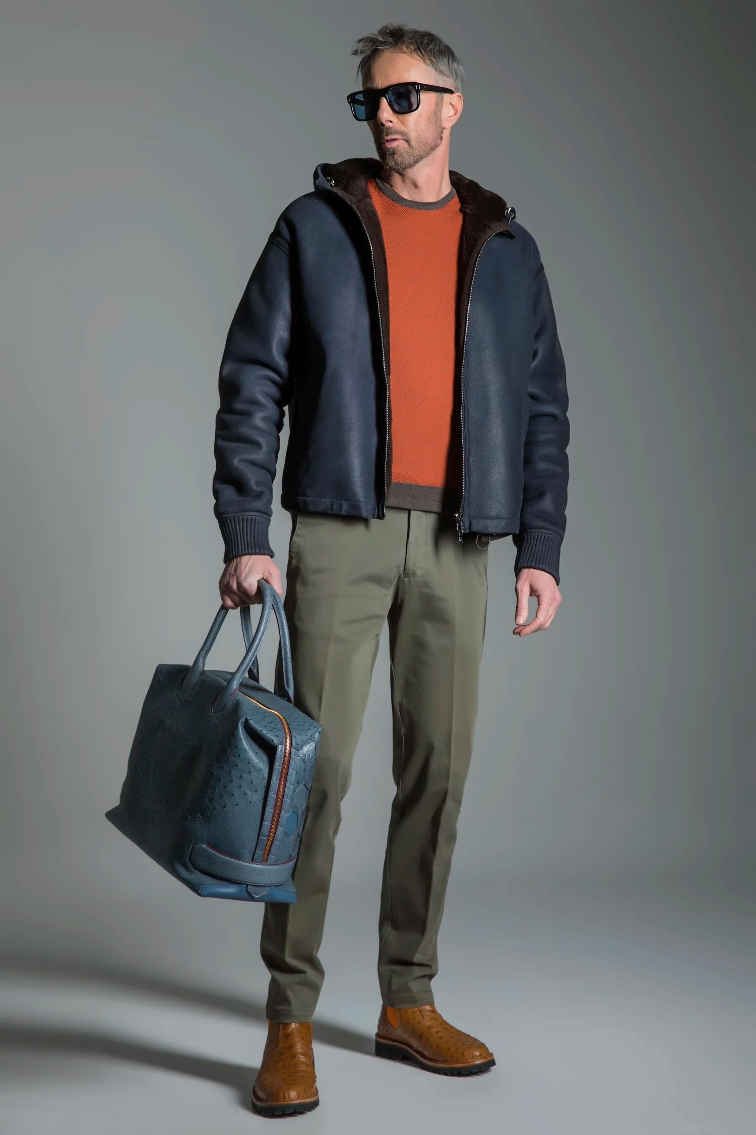 Man wearing a dark jacket, orange sweater, green pants, and brown shoes, holding a blue bag against a gray background.