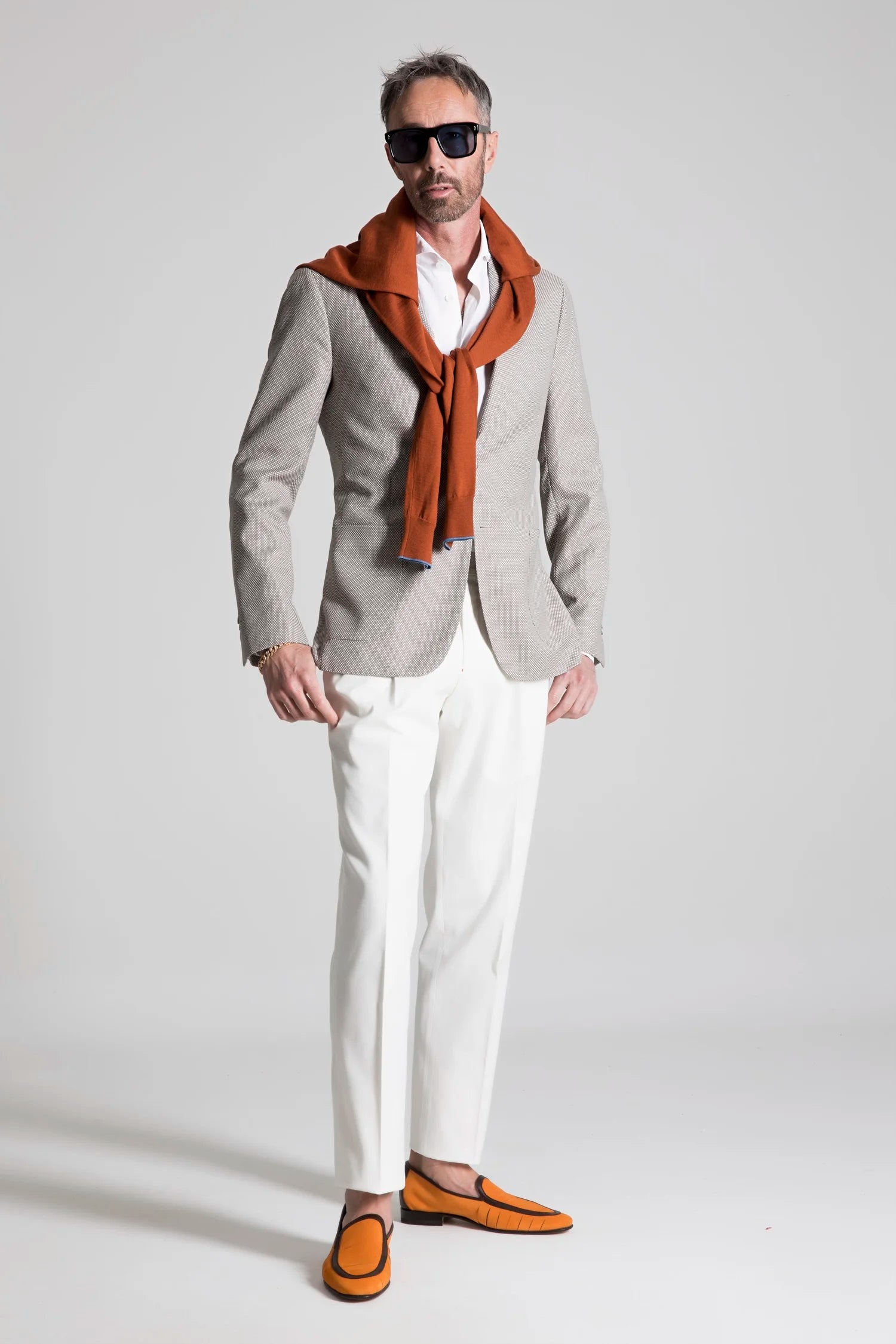 Man wearing a gray blazer, white shirt, white pants, orange shoes, and a rust-colored scarf on a light gray background